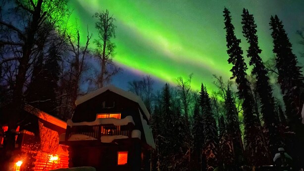 A lodge with a snow-covered roof, surrounded by tall trees, under a night sky with green aurora lights. - ^^LOG CABIN ON THE RIVER! Alaskan Aurora Adventure (Fairbanks)