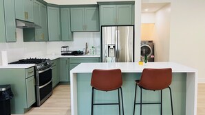 A modern kitchen with green cabinets, a stainless steel refrigerator, and a white countertop. There are two bar stools at the island. - Entire home 3bed w/ Laundry & 2 Spa like baths & private parking w/ EV charging (Long Beach)