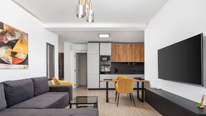 A modern living room with a grey sofa, a small coffee table, and a yellow chair. There is a kitchen area with wooden cabinets and a black countertop. A large flat-screen TV is mounted on the wall. - 1 bedroom apartment A4 in Villa Marealto with pool and garage (Ičići)