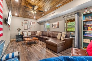A living room with a sectional sofa, wooden flooring, a ceiling fan, and a bookshelf. - Family Camp - Home for 17 (Hollister)