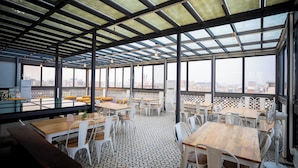 A rooftop dining area with tables and chairs, large windows, and a view of the city. - SHIVA KASHI HOTEL (Varanasi)