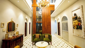 A spacious hallway with a high ceiling, a chandelier, two green armchairs, a round table, and a wooden cabinet. - SHIVA KASHI HOTEL (Varanasi)