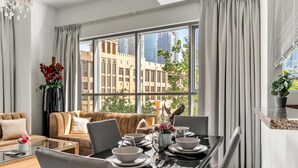 A modern living room with a glass dining table, beige sofas, and a large window with a city view. - StayShort - Downtown 2 Bed Unit with Burj Views that Sleeps 4 (Downtown)