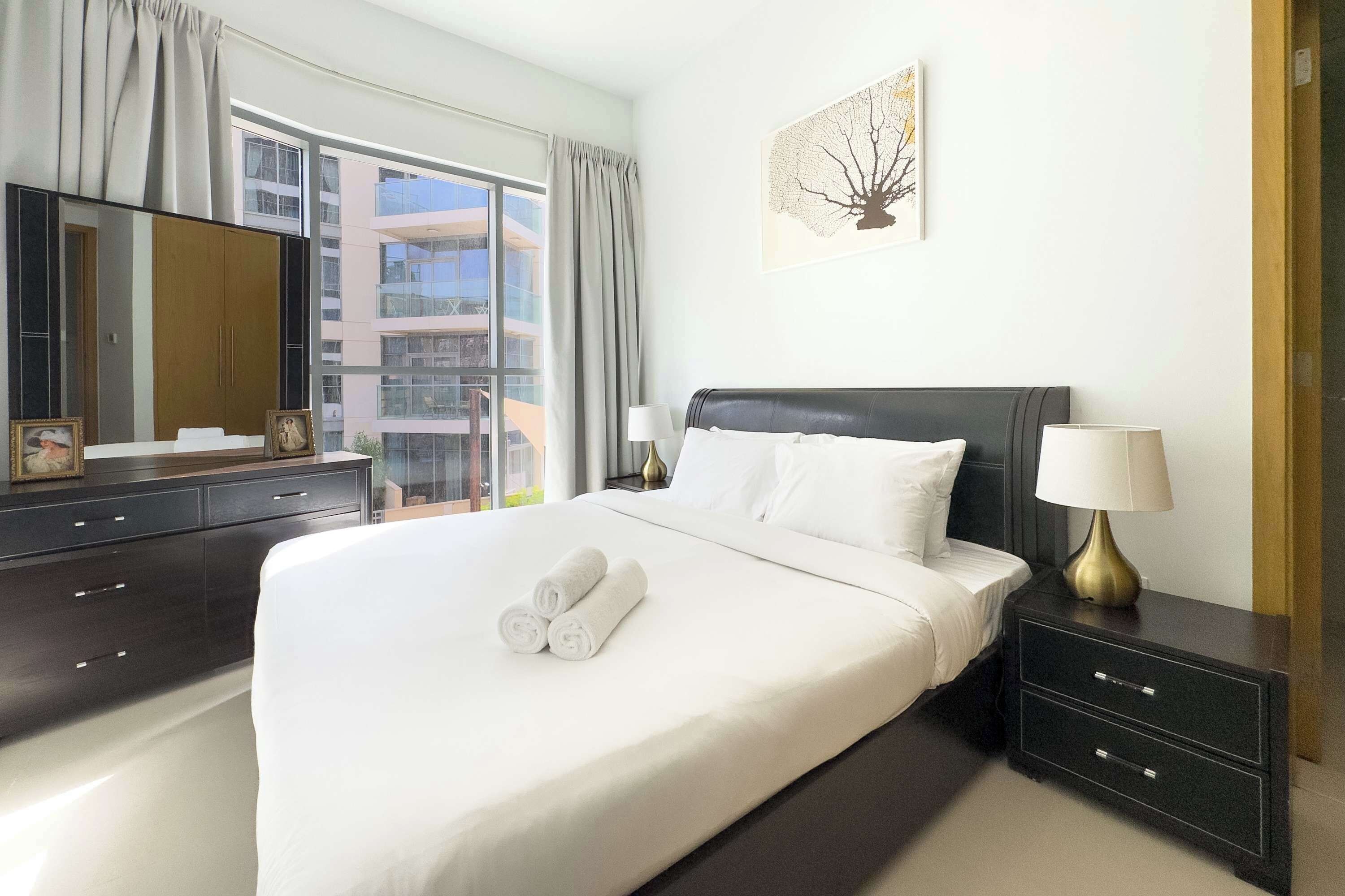 Una habitación de hotel con una cama grande, una mesita de noche oscura con lámpara, un tocador con espejo y una ventana con cortinas.