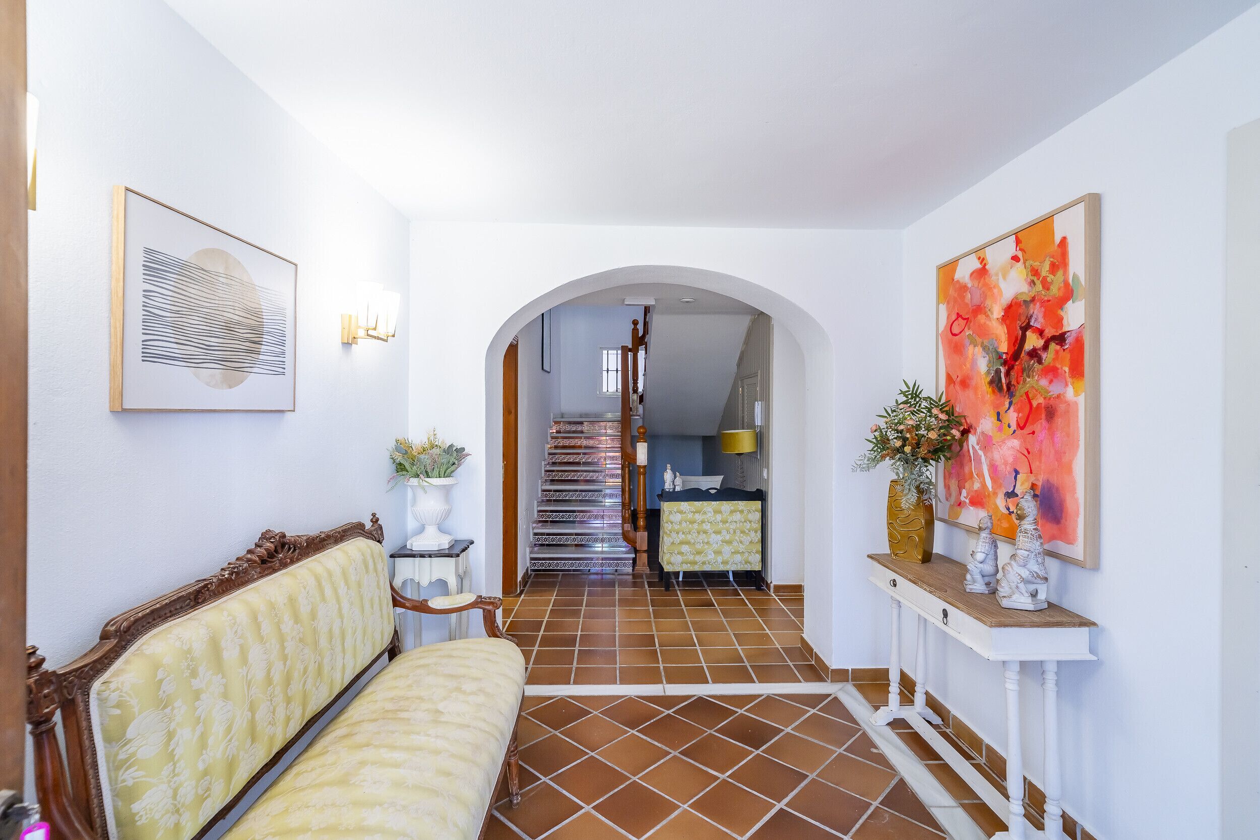 A hallway with a sofa, a console table with decorative items, and a staircase leading to a kitchen area.