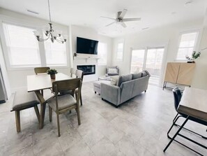 A spacious living room with a gray sofa, a dining table with chairs, a chandelier, and a ceiling fan. - Beach Block heart of Downtown Walk to Everything (Sea Isle City)