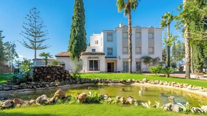 A white building with a tiled roof, a stone wall, and a pond with rocks and plants. - Cubo’s NAZ Country Stay | Single Room with Shared Bathroom + Optional Breakfast (Alhaurín el Grande)