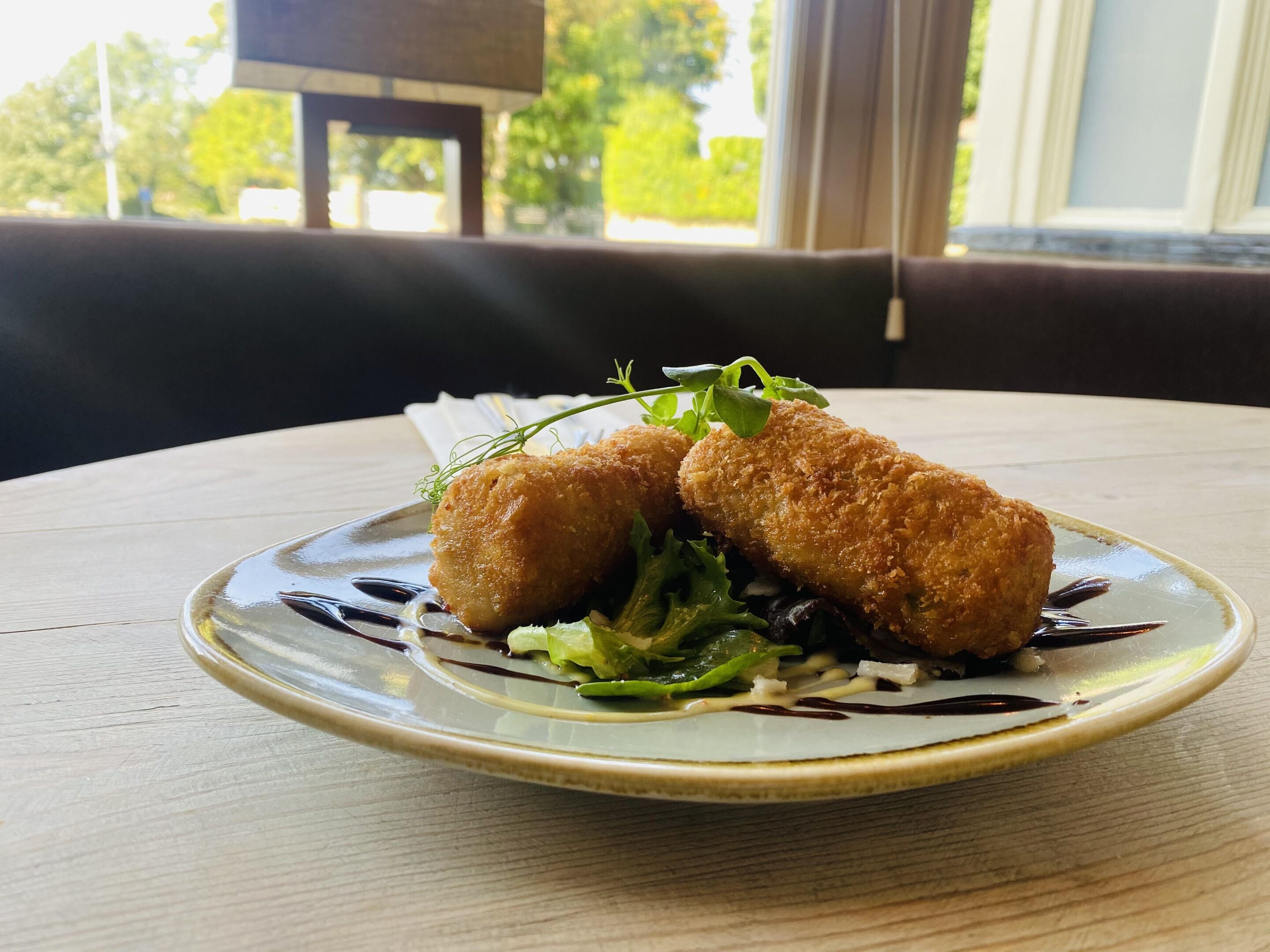 Un plato con dos croquetas doradas, ensalada verde y una salsa oscura sobre un plato de cerámica.