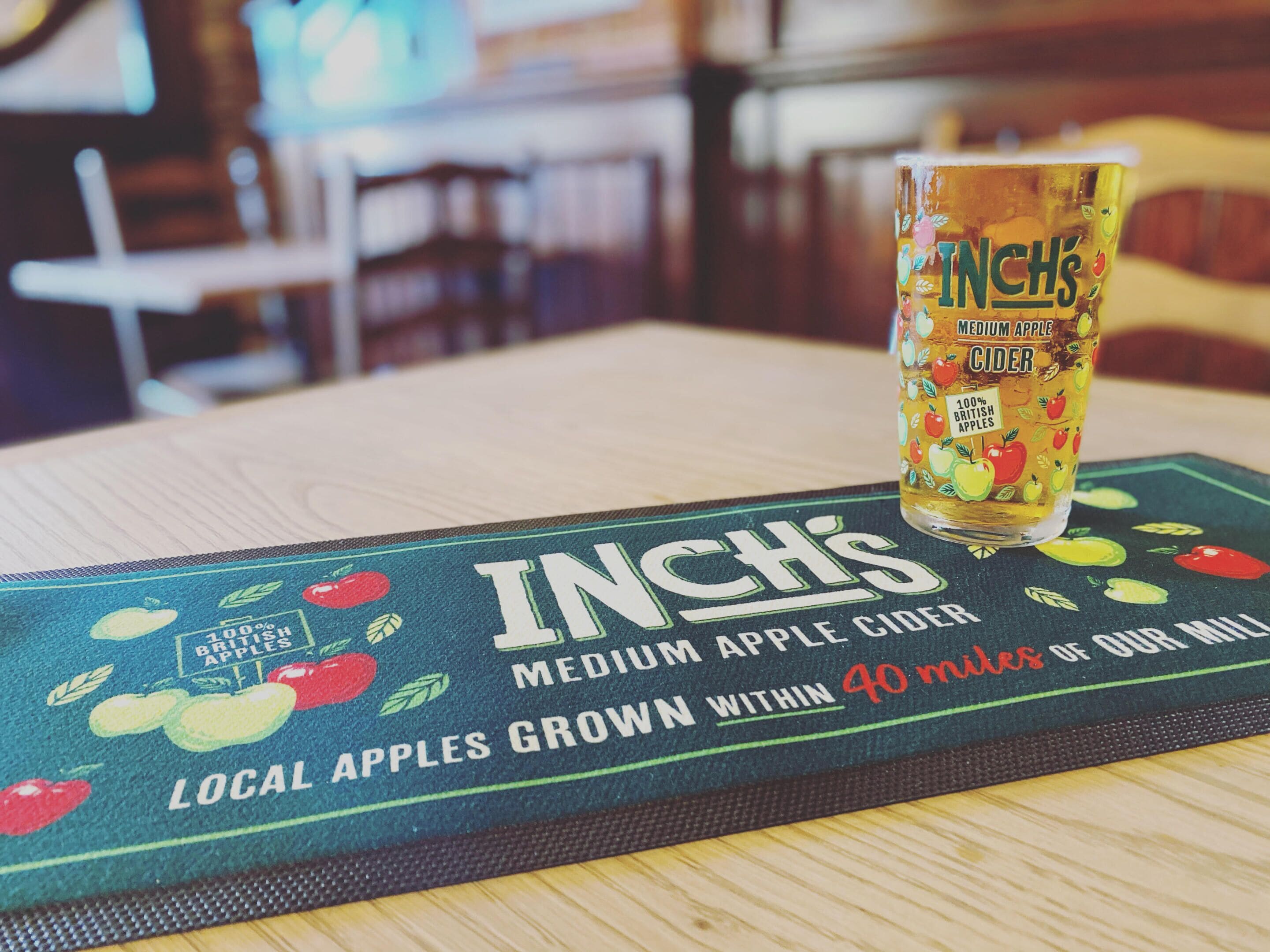Un vaso de sidra de manzana mediana de Inch sobre una mesa con un mantel individual que presenta ilustraciones de manzanas y texto sobre el abastecimiento local de manzanas.