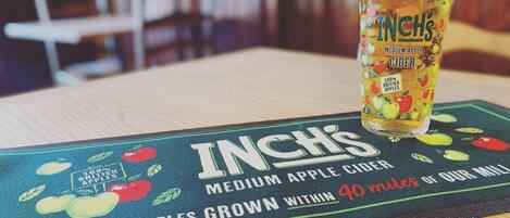 Un vaso de sidra de manzana mediana de Inch sobre una mesa con un mantel individual que presenta ilustraciones de manzanas y texto sobre el abastecimiento local de manzanas.