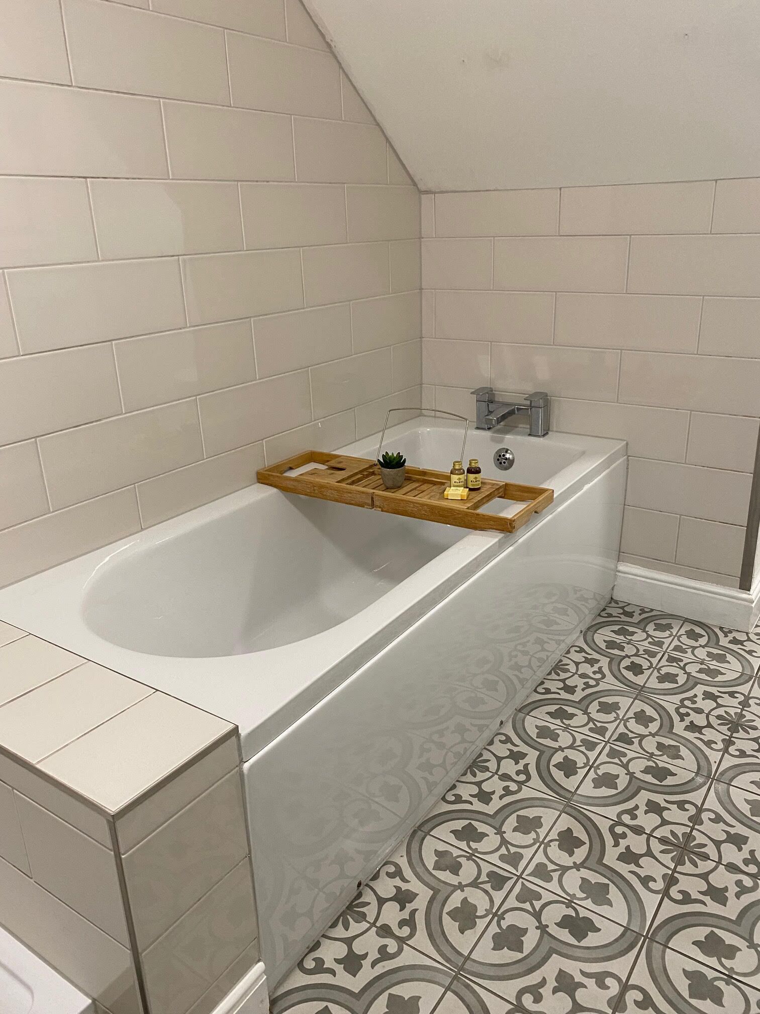 A modern bathroom with a freestanding bathtub, a wooden tray with toiletries, and patterned floor tiles.