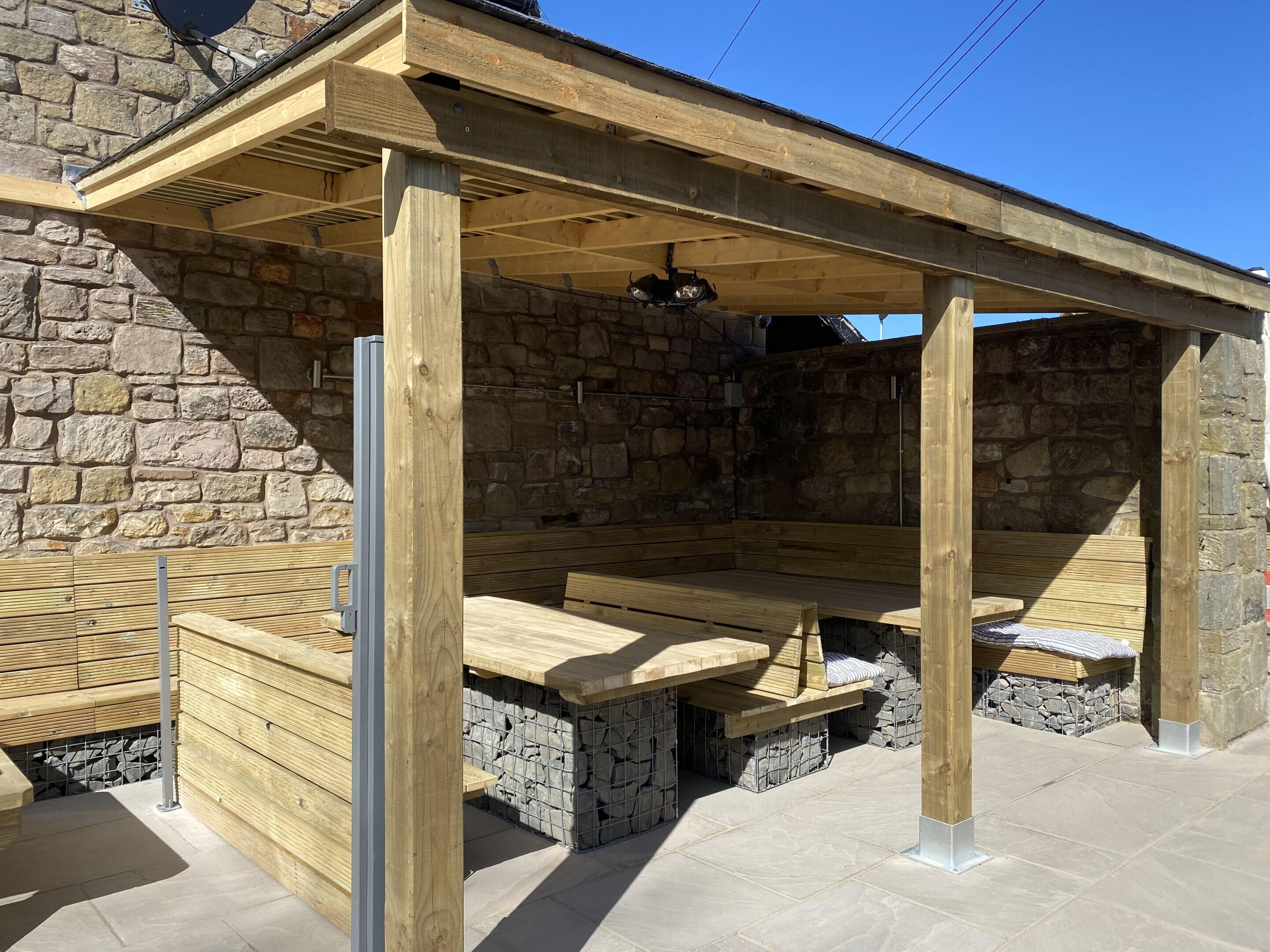 A wooden pergola structure with benches, stone walls, and a clear blue sky.