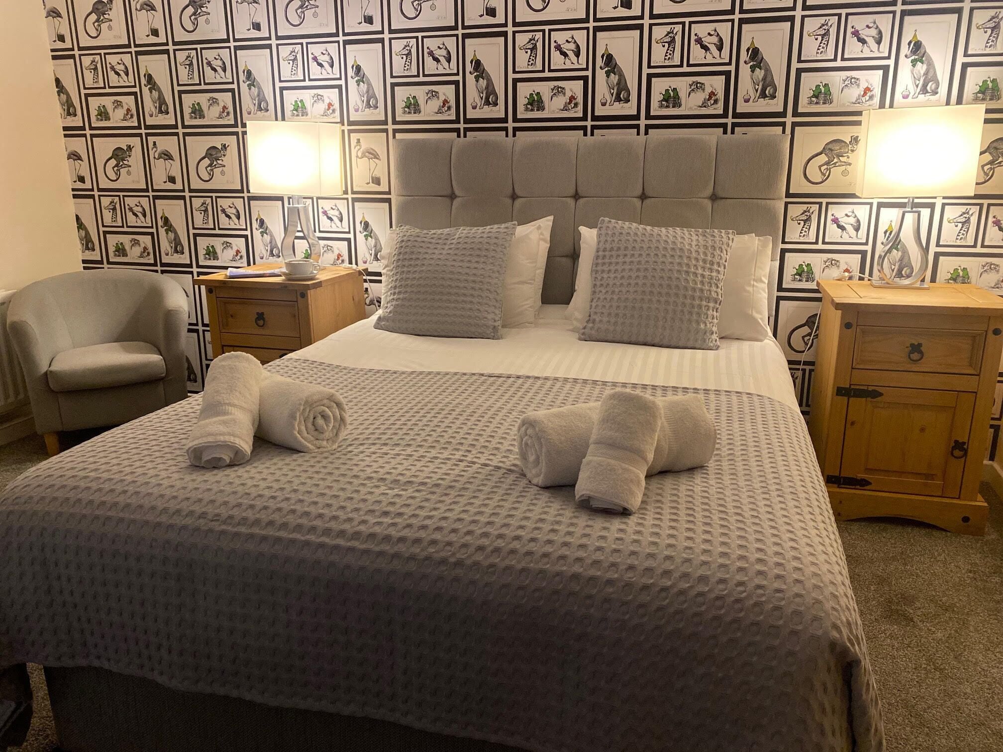 A neatly made bed with a grey textured bedspread and pillows, two rolled towels on the bed, a grey armchair, and a nightstand with a lamp.