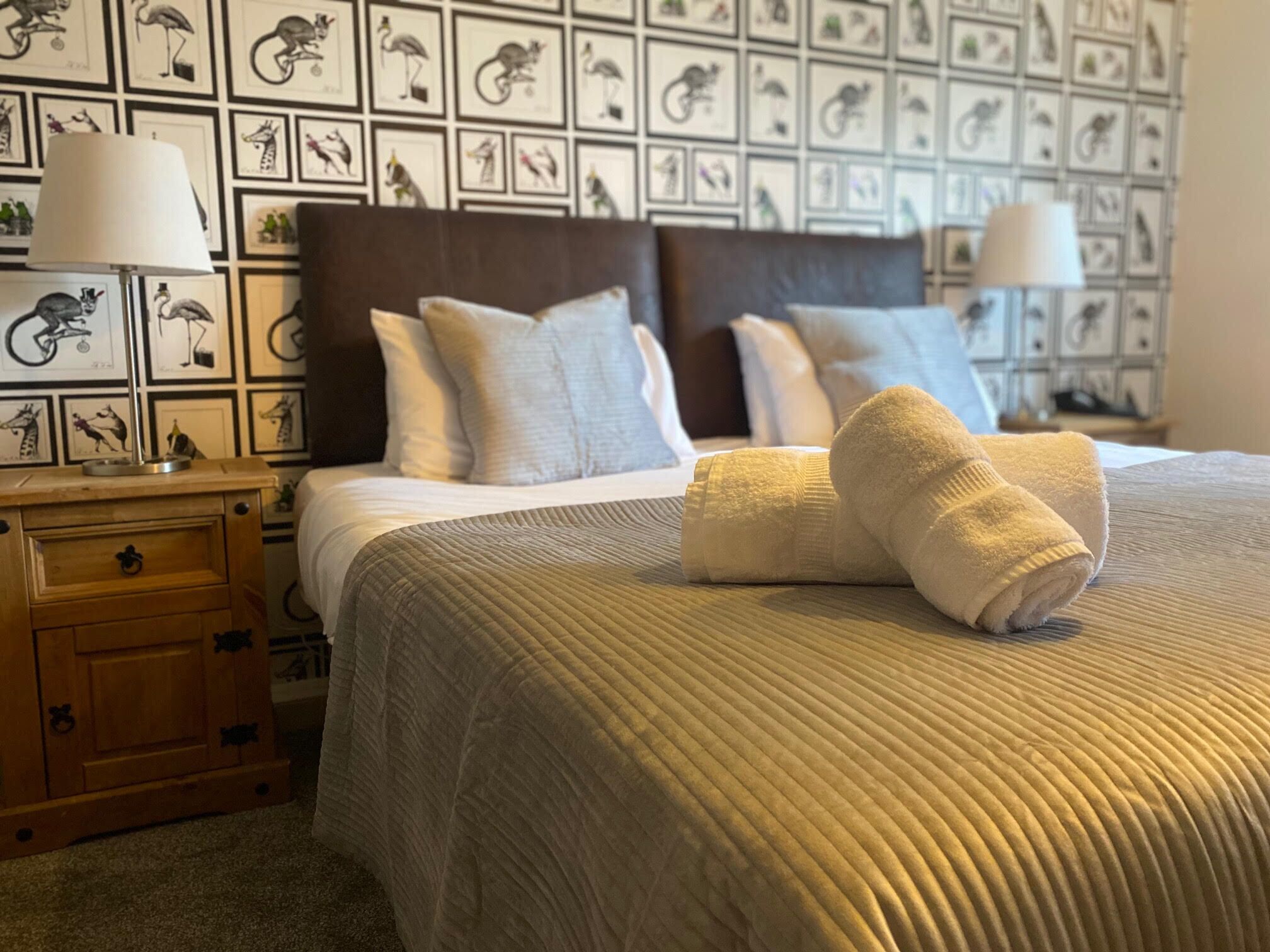 Ein ordentlich bezogenes Bett mit einer beigen Steudecke, zwei Kissen und einem zusammengerollten Handtuch auf einem Nachttisch aus Holz.
