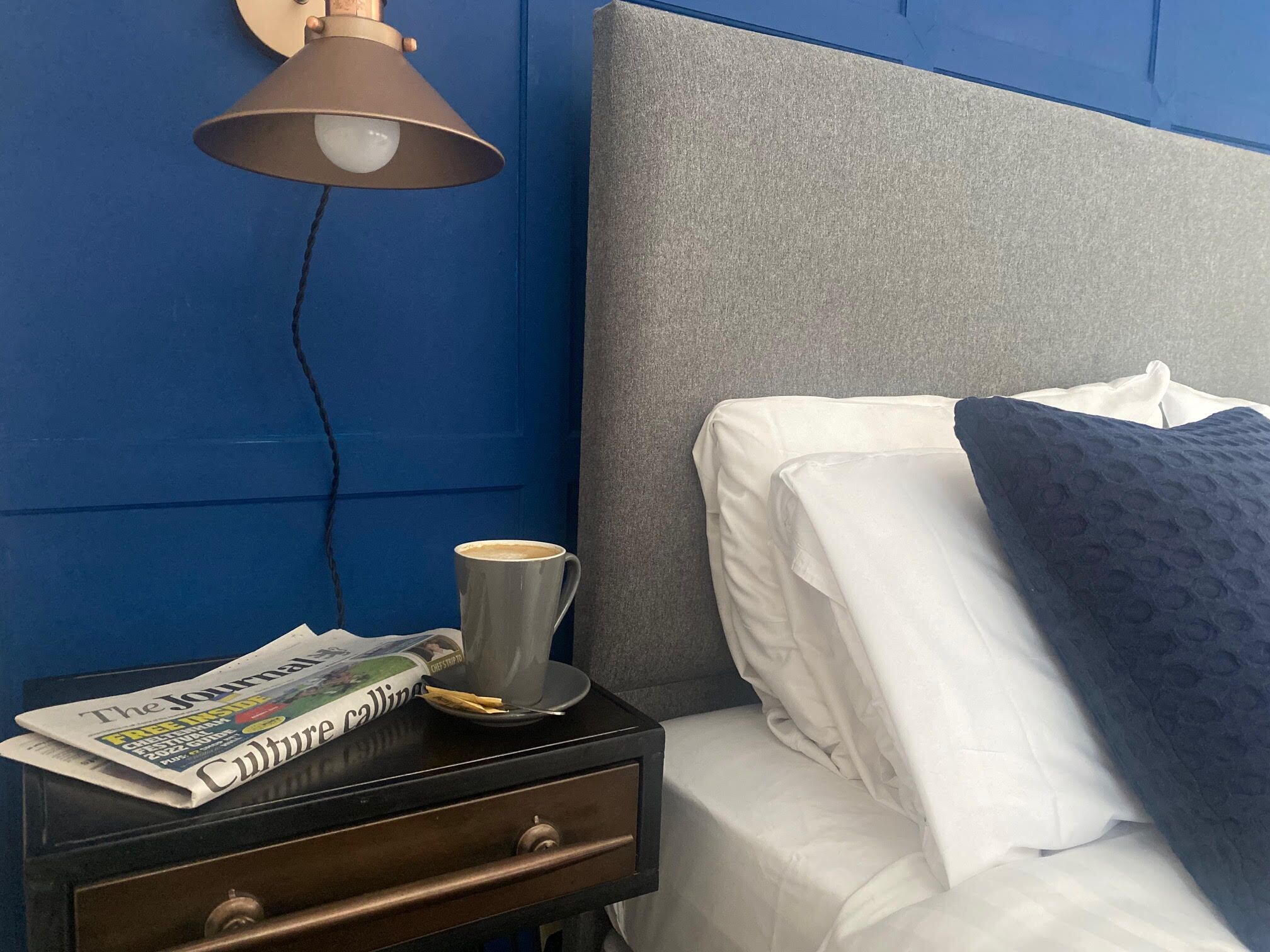 A neatly made bed with white linens and a dark blue pillow, a bedside table with a lamp, a cup of coffee, and a newspaper.
