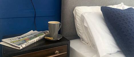 A neatly made bed with white linens and a dark blue pillow, a bedside table with a lamp, a cup of coffee, and a newspaper.