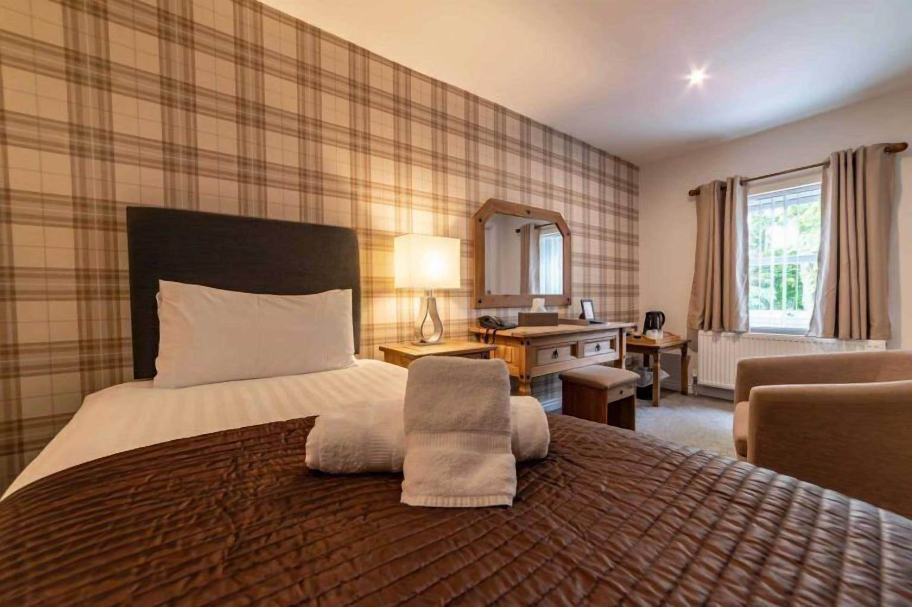 Habitación de hotel con una cama grande, una mesita de noche con lámpara, cómoda, espejo y ventana con cortinas.