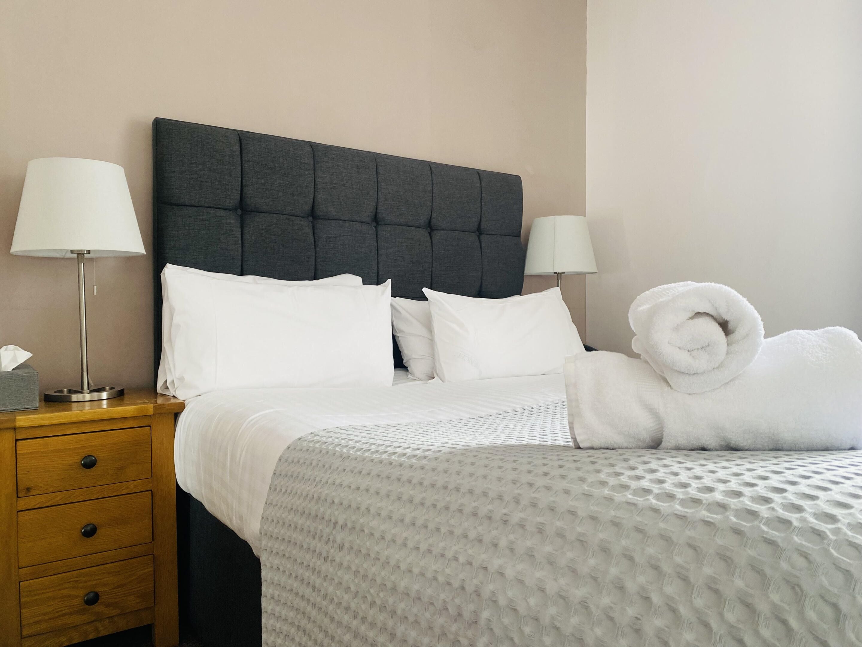 A neatly made bed with white linens and a grey textured blanket, flanked by two bedside lamps and a wooden nightstand with drawers.