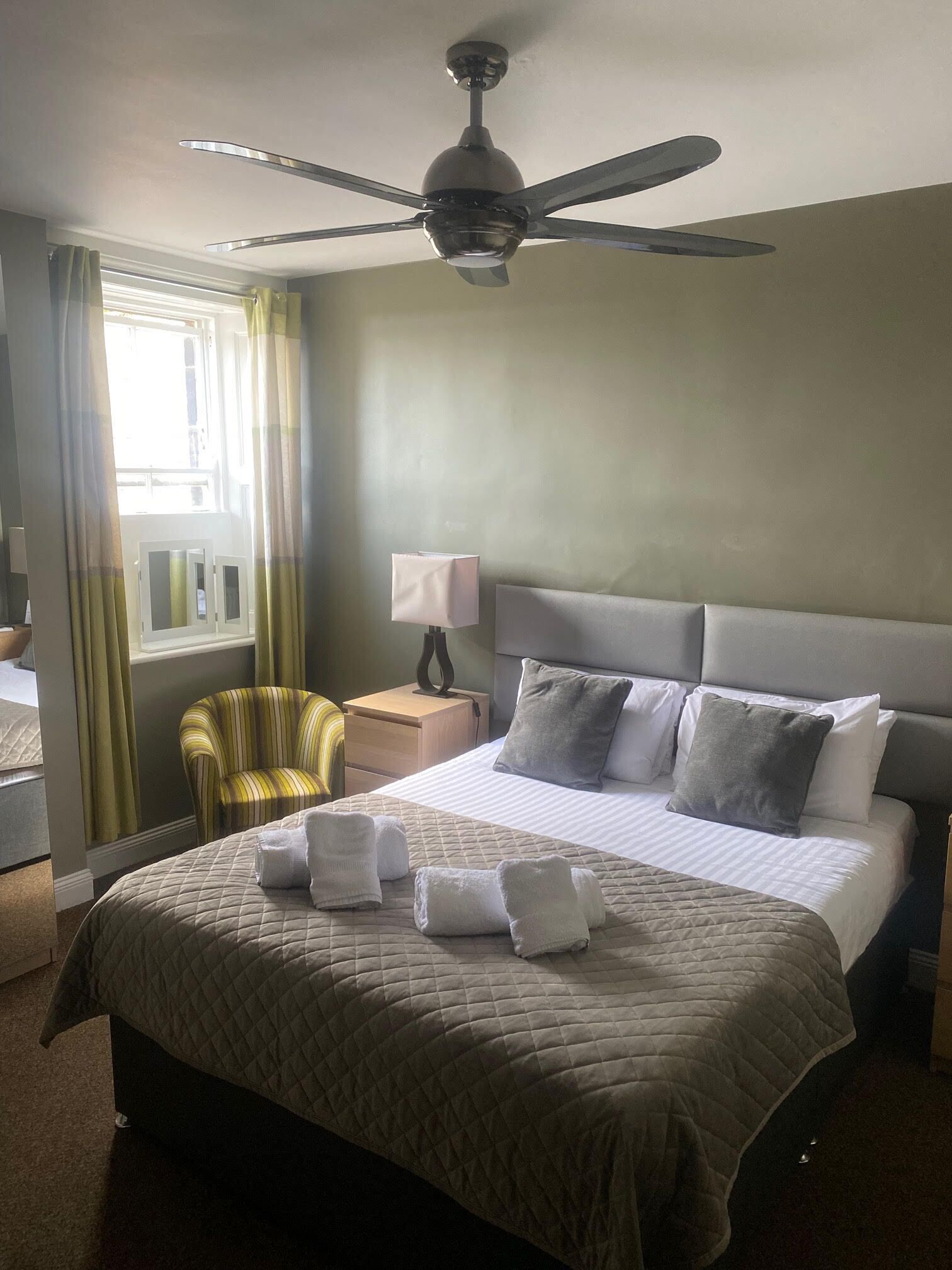 A hotel room with a bed, a chair, a nightstand, a ceiling fan, and a window with curtains.