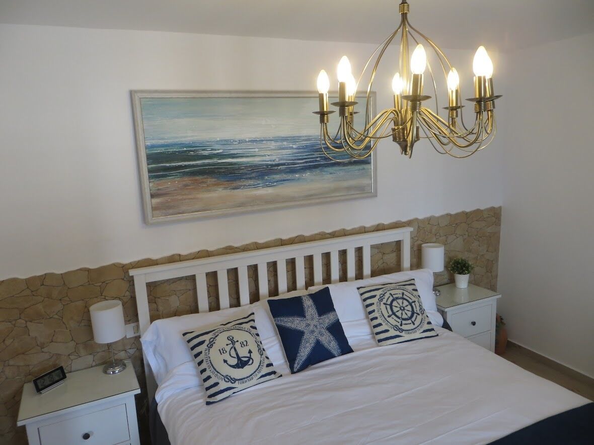 A bedroom with a bed, a nightstand, a lamp, a wall-mounted painting, and a chandelier.