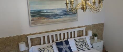 A bedroom with a bed, a nightstand, a lamp, a wall-mounted painting, and a chandelier.