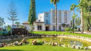 A white building with a tiled roof, a stone wall, and a pond with rocks and plants.