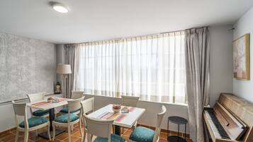 A room with a piano, a dining table with chairs, a statue, and a window with curtains.