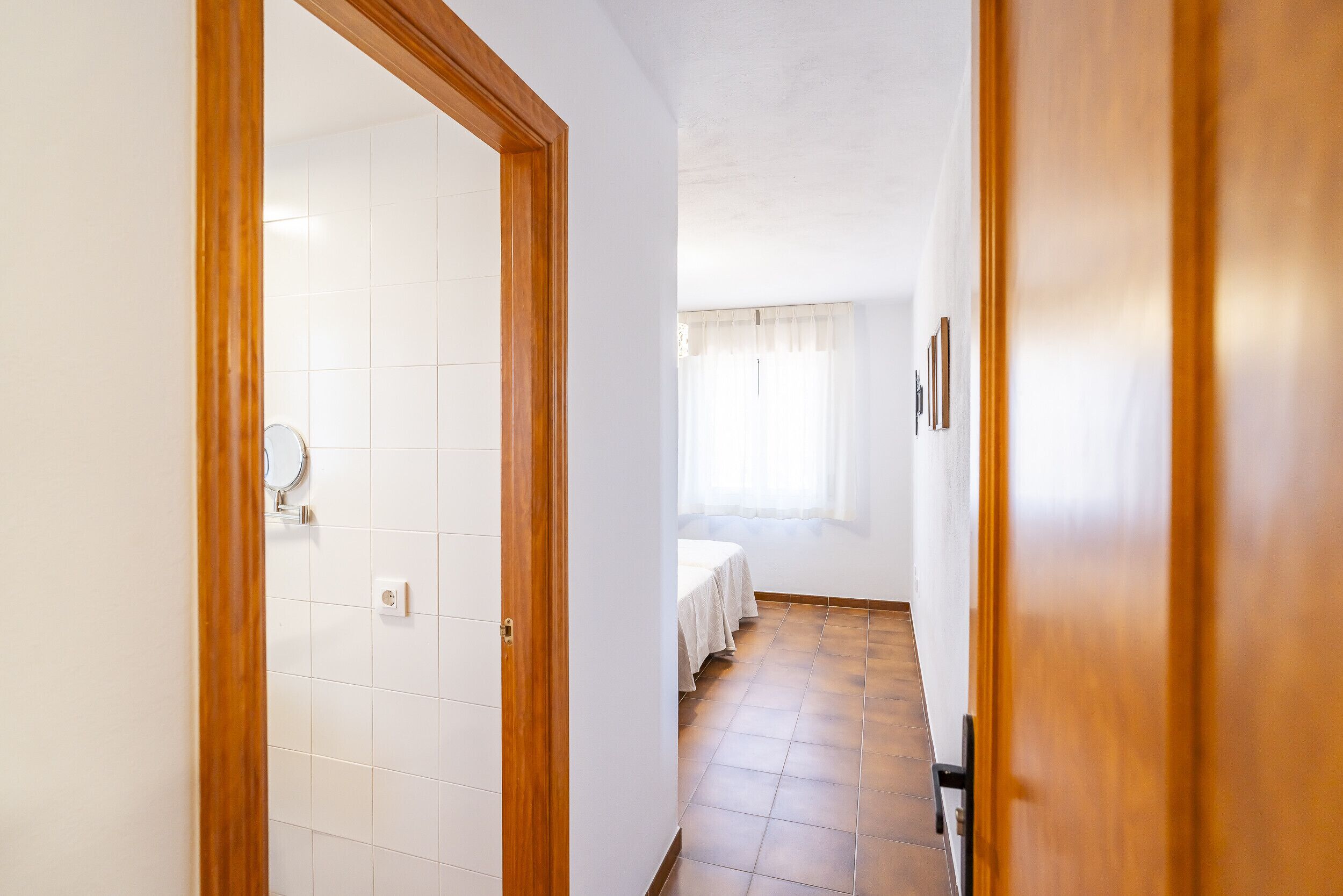 A hallway with a wooden door frame, white tiled walls, a bed with a white bedspread, and a window with sheer curtains.
