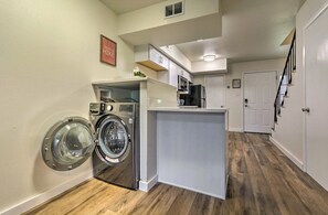 Een functionele wasruimte met een wasmachine en droger, een klein keukenhoekje met een koelkast, en een deur naar een andere kamer.