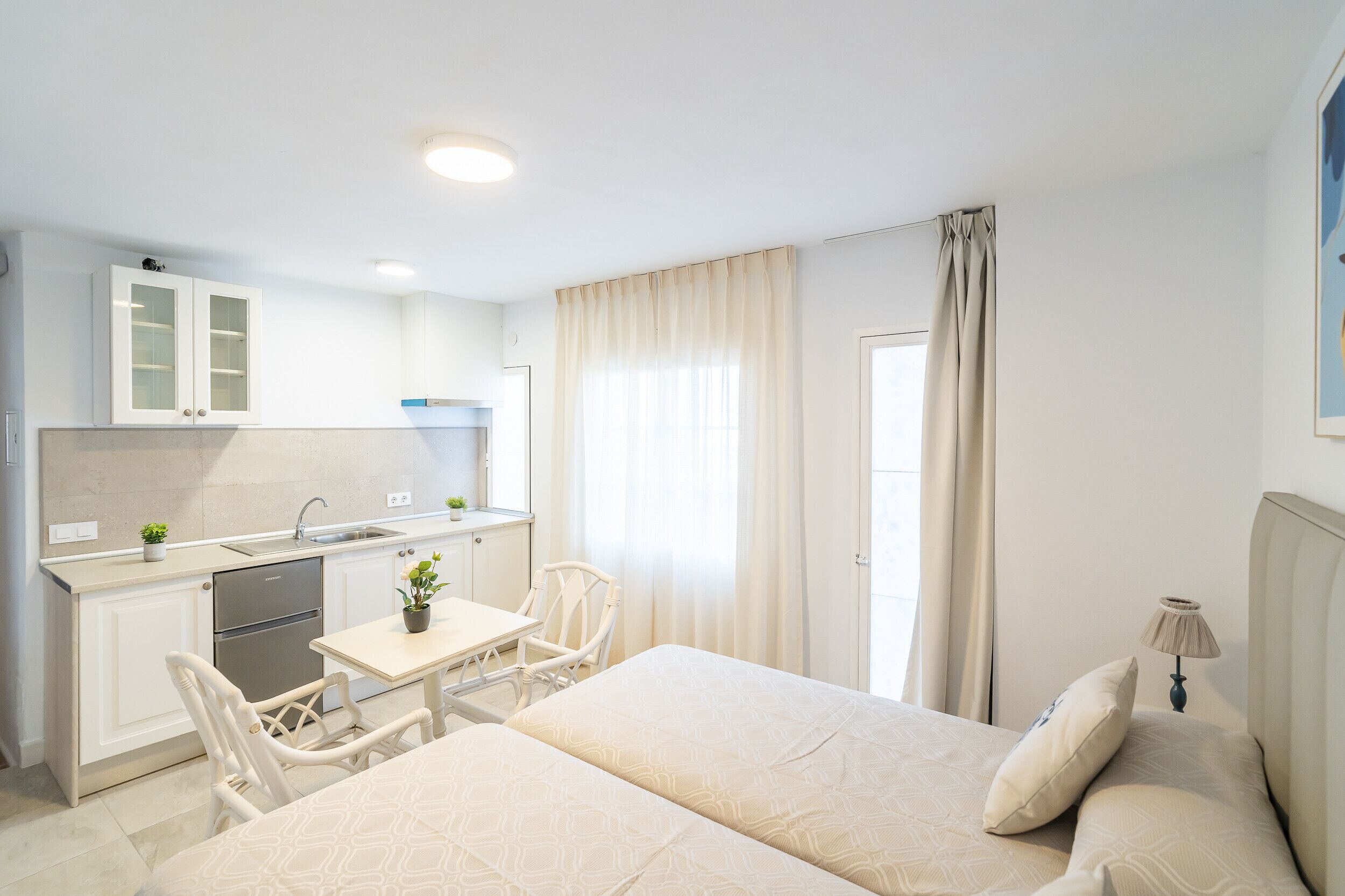 Habitación de hotel compacta con kitchenette, cama con dos almohadas, una mesita y una silla.