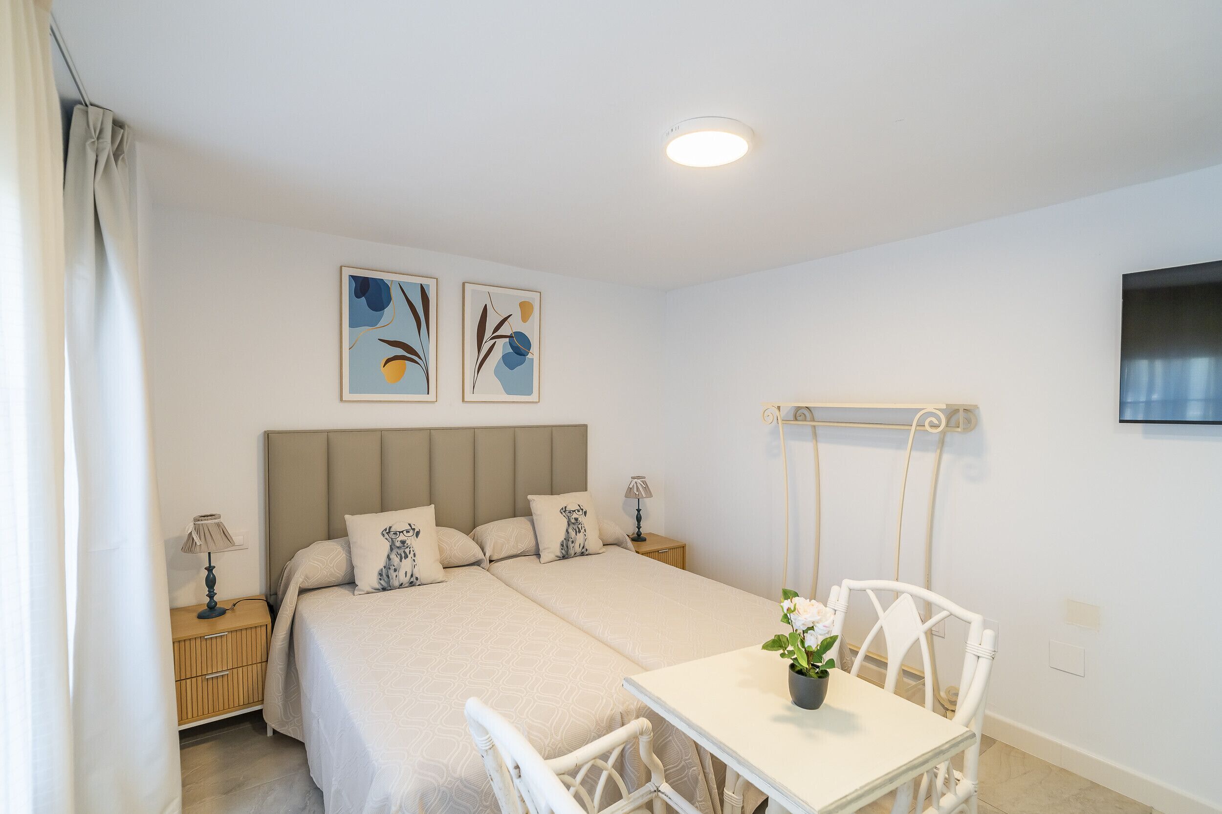 A double bed with a headboard, two bedside tables with lamps, a small table with a chair, and two abstract wall art pieces.