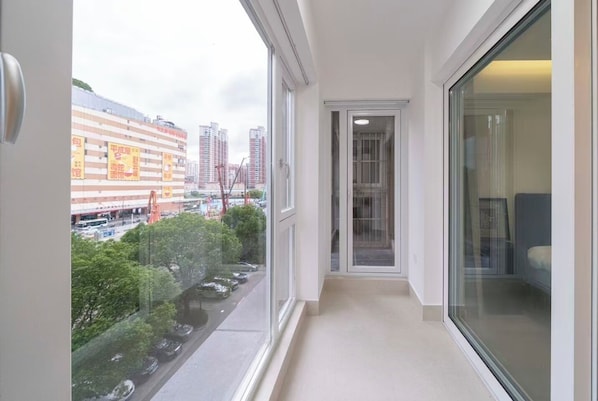 Un corridoio stretto con una porta in vetro che dà su una stanza, una vista sulla città con edifici e una strada, e un balcone affacciato sulla strada e sul verde.