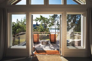 A room with large windows, a wooden deck, outdoor seating, and a view of trees and a body of water.