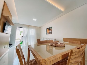 A modern living room with a glass-top dining table, wicker furniture, a painting on the wall, and a TV mounted on the wall. - Residencial Jardins de Taperapuã 2 (Porto Seguro)