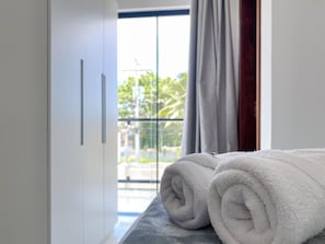 A neatly made bed with a rolled white towel, a window with a view of greenery, and a door with a handle. - Residencial Jardins de Taperapuã 2 (Porto Seguro)
