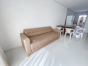 A modern living room with a beige sofa, a wooden dining table, and white plastic chairs. - Residencial Jardins de Taperapuã 2 (Porto Seguro)