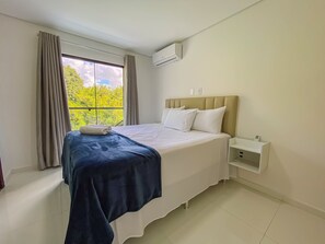 A hotel room with a bed, a bedside table, an air conditioning unit, and a window with curtains. - Residencial Jardins de Taperapuã 2 (Porto Seguro)