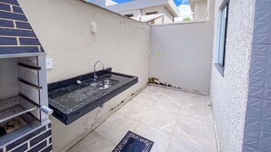 A small outdoor kitchen with a sink and a grill, a tiled floor, and a white wall with a window.