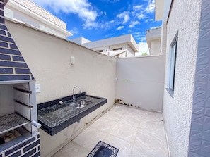 Una pequeña cocina al aire libre con un lavamanos y una parrilla, suelo de baldosas y una pared blanca con una ventana.
