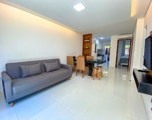 Sala de estar com sofá, mesa de jantar, cadeiras, televisão e área de cozinha ao fundo.