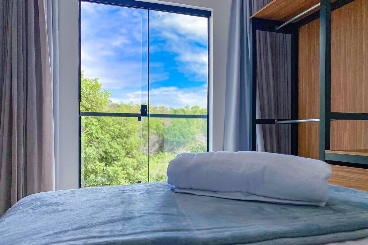 A bed with a white pillow, a window with curtains, and a view of greenery outside.