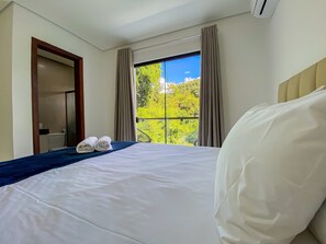 A hotel room with a bed, a window with curtains, a view of greenery, and an air conditioning unit. - Residencial Jardins de Taperapuã 2 (Porto Seguro)