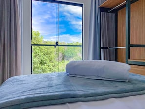 Una cama con un almohadón blanco, una ventana con cortinas y vistas al verdor exterior.