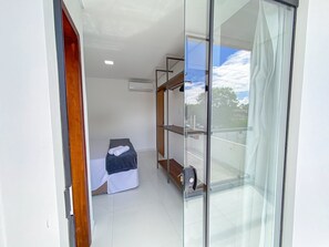 A small, modern room with a glass door leading to a balcony, a bed with a dark cover, and a shelf unit with various items.