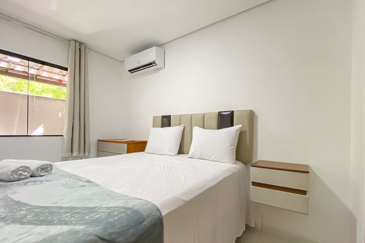A hotel room with a bed, a bedside table, a nightstand, a window with curtains, and an air conditioning unit.