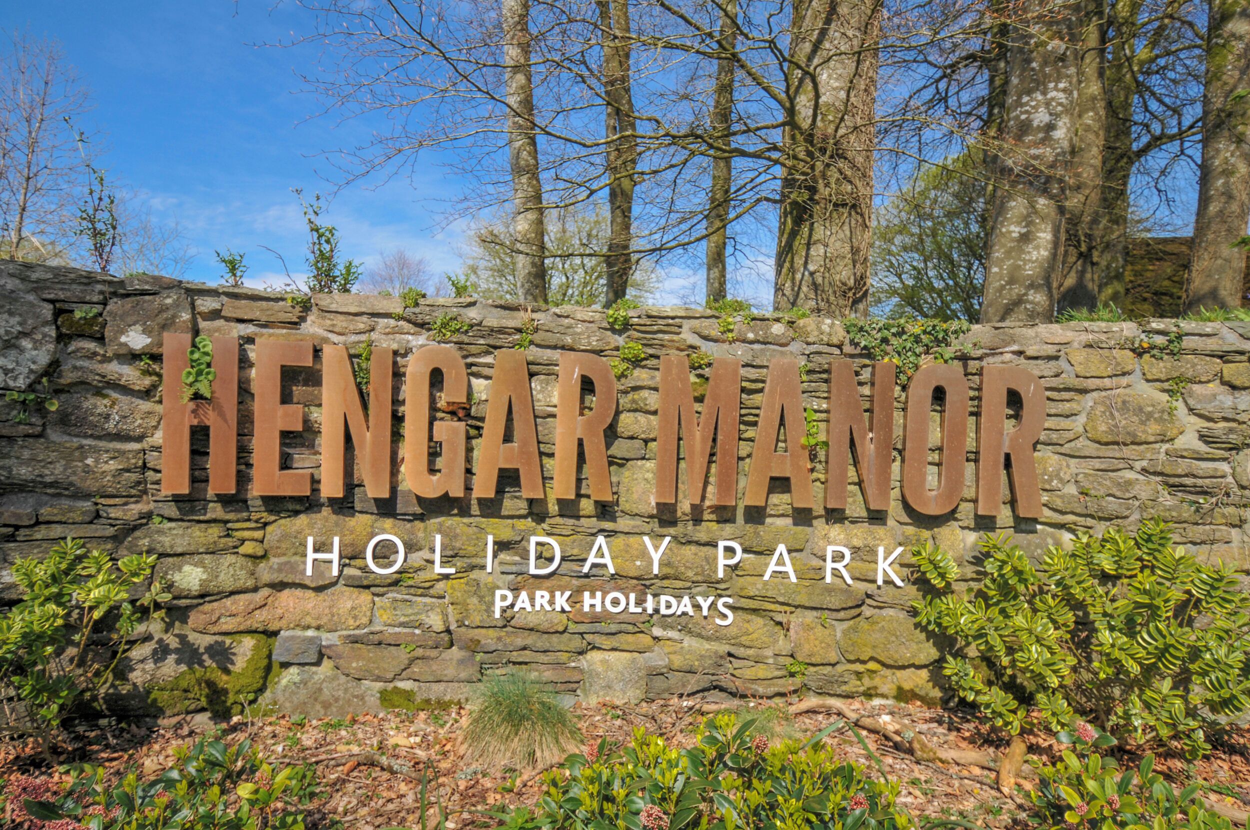 A stone wall with 'Hengar Manor Holiday Park' signage, surrounded by greenery and trees.