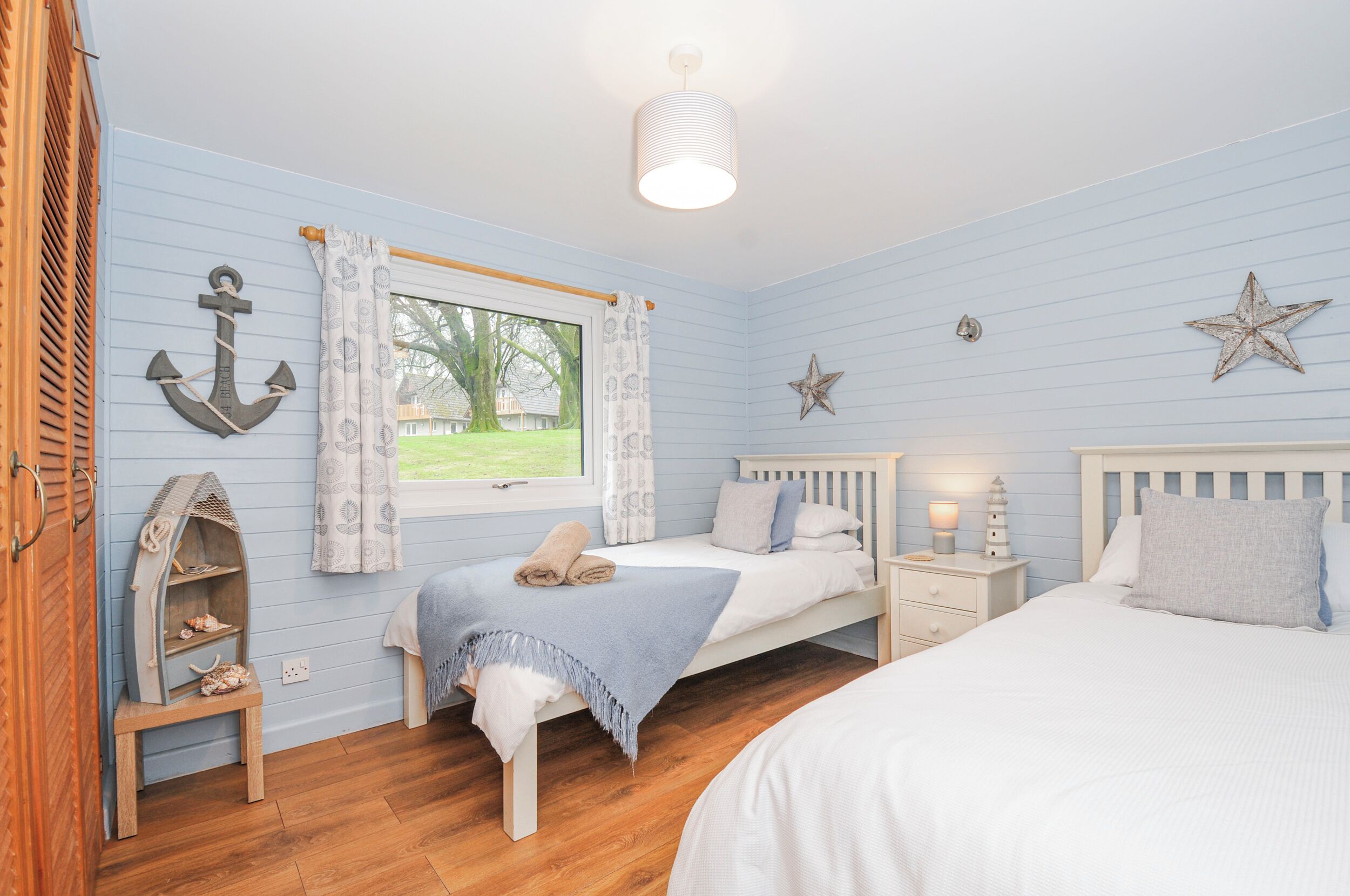 A bedroom with two beds, a window with curtains, a wooden floor, and nautical-themed decor.