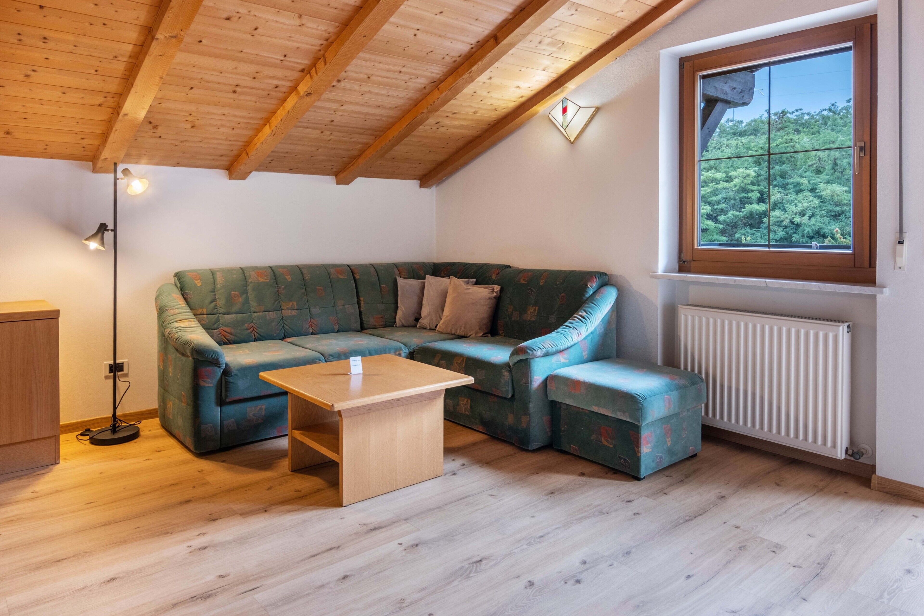 Een kamer met een houten plafond, een groene bank, een houten tafel, een raam met uitzicht op bomen en een radiator.