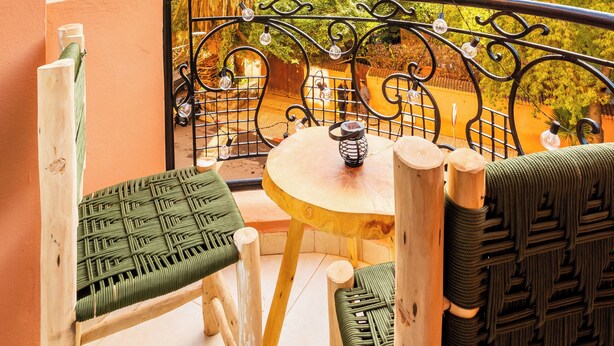 Uma varanda com vista para palmeiras e edifícios, uma mesa redonda com cadeiras, e um guarda-corpo de metal decorativo.