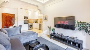 Sala de estar moderna com um sofá cinzento, uma televisão de tela plana e uma cozinha ao fundo.